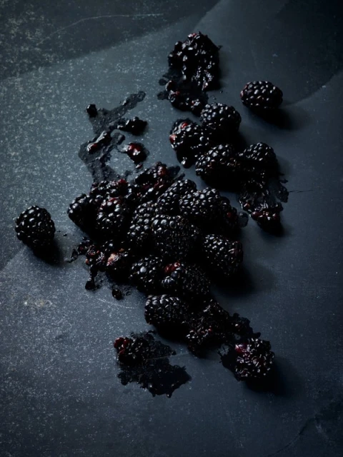 Overhead shot of fresh blackberries spread across a dark gray stone surface, some crushed, creating a rich, dark and textured composition.