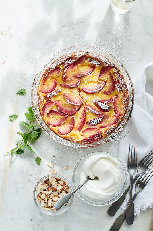 Overhead shot of plum clafoutis in glass baking dish with powdered sugar, mint sprigs, whipped cream, sliced almonds, and forks.