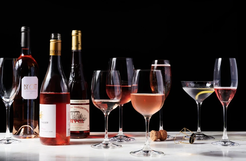 Arrangement of rosé wine bottles and filled wine glasses on a white marble surface against a dark background in a studio setting.