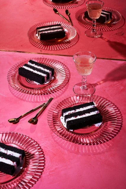 Slices of black and white cake on clear glass plates with gold spoons and champagne flutes are arranged on a pink surface.