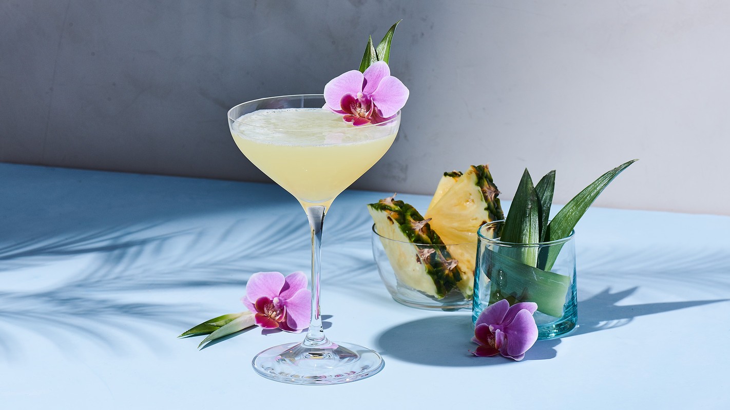 Elegant cocktail drink in stemmed glass, garnished with an orchid, next to pineapple slices and greenery, blue background with shadows.