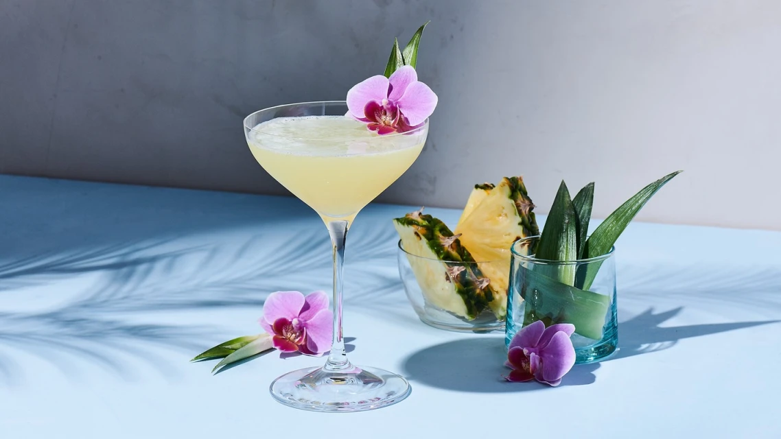 Elegant cocktail drink in stemmed glass, garnished with an orchid, next to pineapple slices and greenery, blue background with shadows.