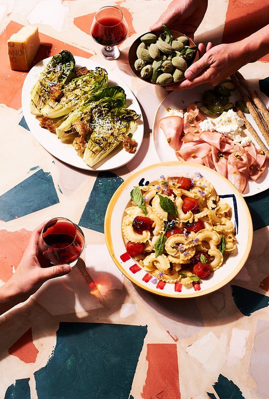 Overhead shot of a table with an assortment of food plates including pasta, lettuce salad, charcuterie, almonds and red wine.