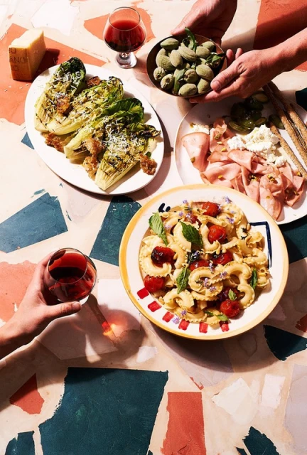 Overhead shot of a table with an assortment of food plates including pasta, lettuce salad, charcuterie, almonds and red wine.