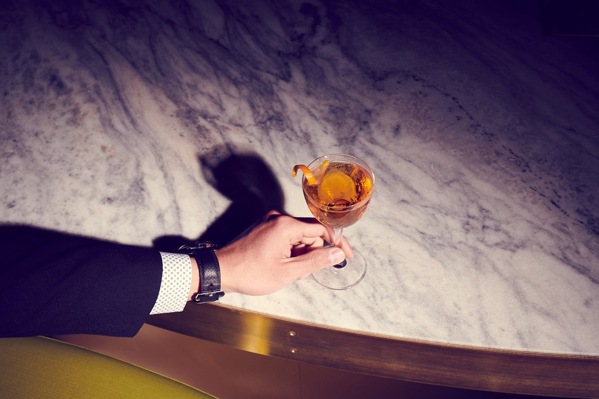 An arm in a suit holds an amber cocktail with an orange twist over a light marble table with gold trim in a dark moody setting.