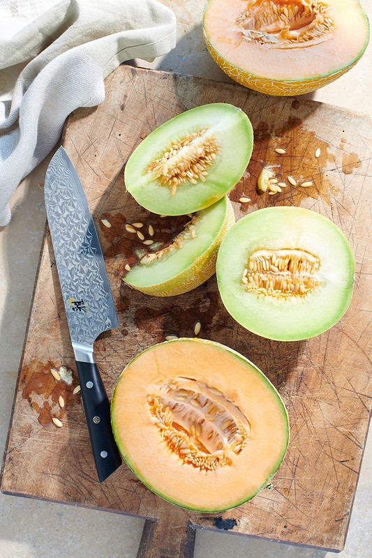 Overhead shot of cantaloupe and honeydew melon halves and slices arranged on a wood cutting board, along with a chef's knife.