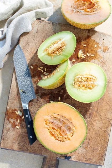 Overhead shot of cantaloupe and honeydew melon halves and slices arranged on a wood cutting board, along with a chef's knife.