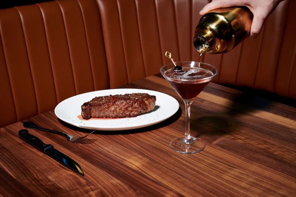 Elevated shot of cooked steak on a plate, next to a cocktail glass being poured with a drink, set on a wooden table with a leather booth.