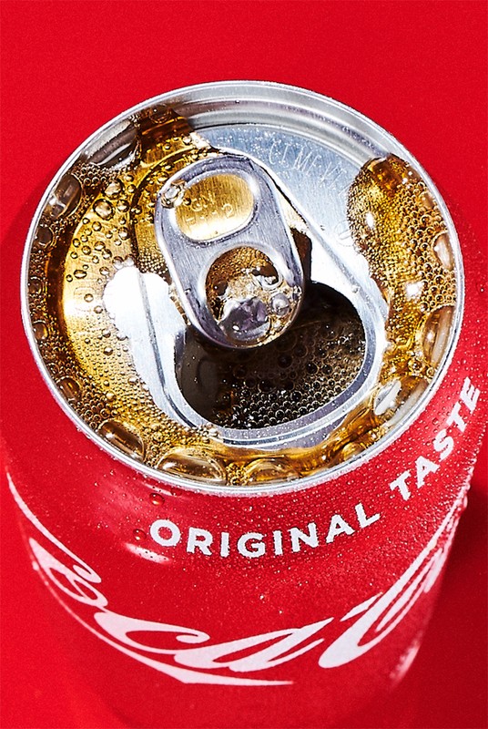 Close-up studio shot of an open Coca-Cola can with water droplets on a red backdrop, showcasing the drink and its condensation.
