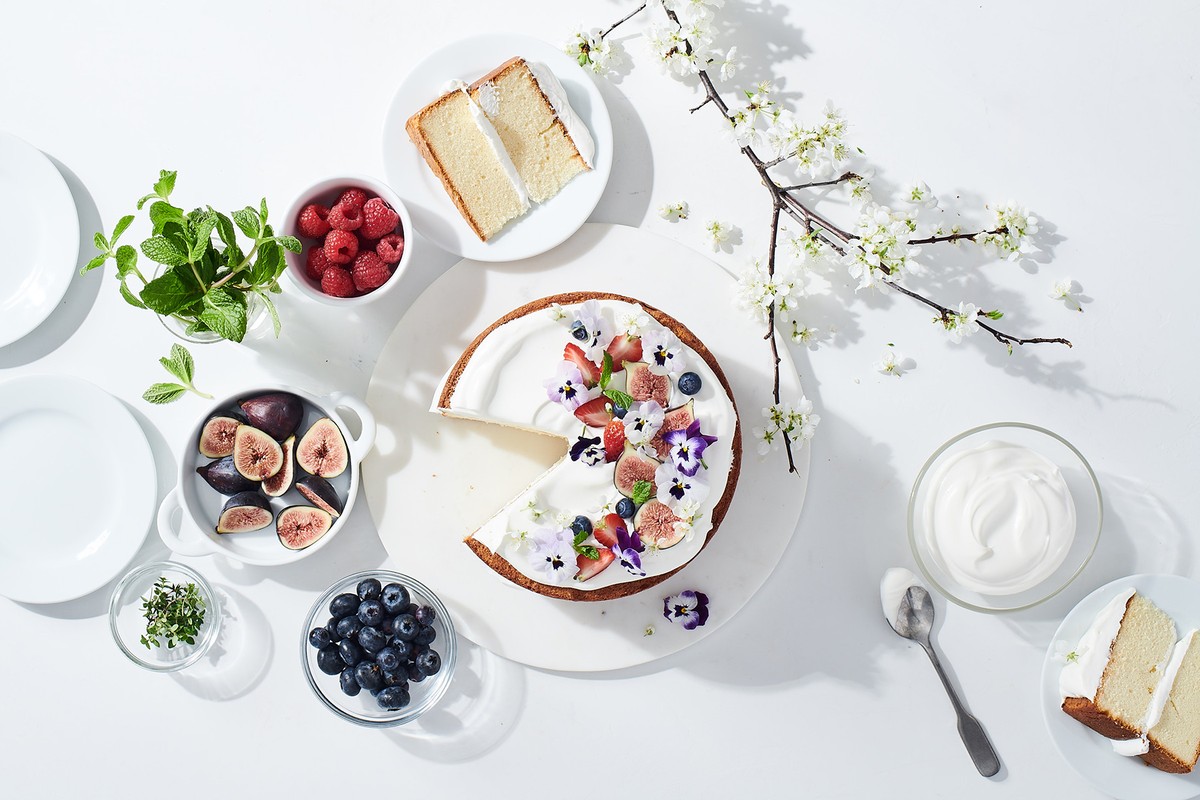 Overhead shot of a frosted cake with fresh fruit and edible flowers, surrounded by bowls of blueberries, figs, raspberries, and mint.