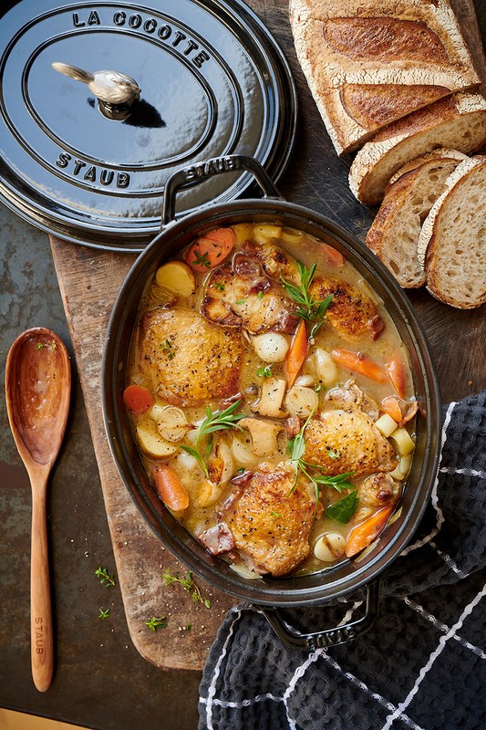 Overhead shot of Staub cocotte with chicken stew, featuring golden-brown chicken thighs, carrots, potatoes, mushrooms, and fresh herbs.