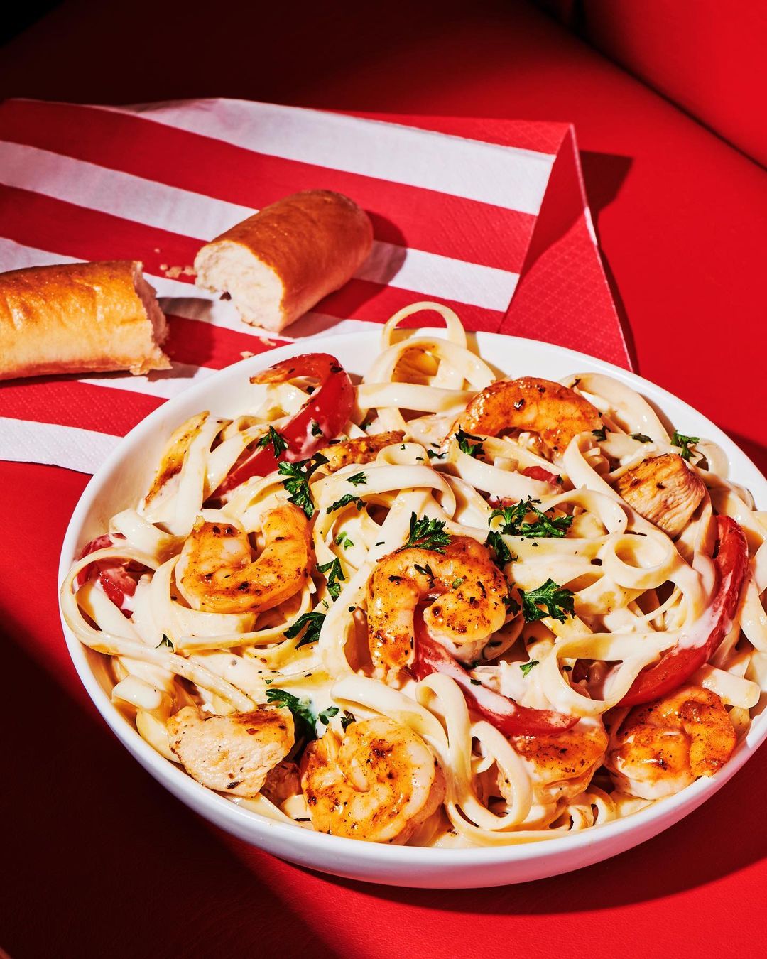 Overhead shot of linguine pasta with grilled shrimp and sliced tomatoes in a white bowl with bread, set on a red striped napkin.