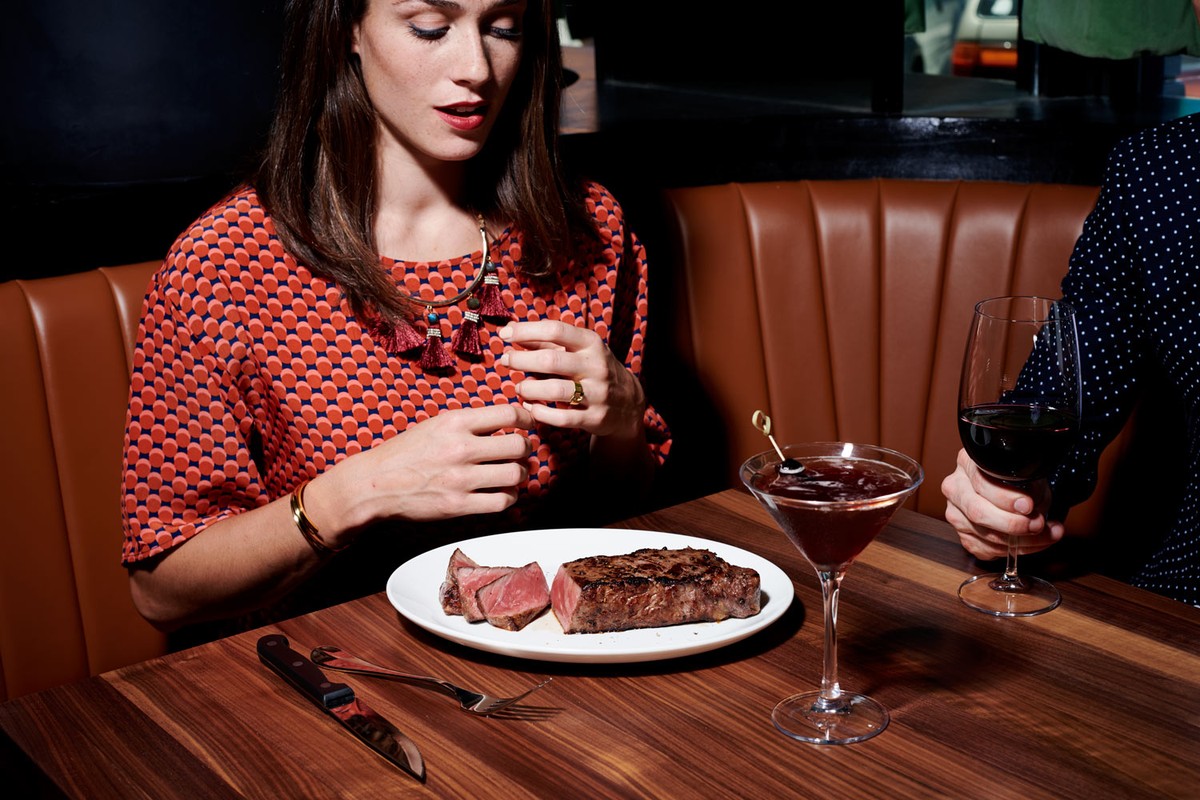 Woman in a patterned shirt at a dimly lit restaurant. A plate with steak and a cocktail glass sit on the table with wine nearby.