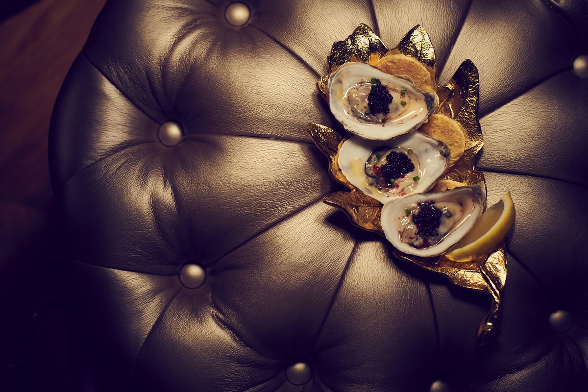 Close-up of three oysters topped with caviar, served on a gold leaf-shaped tray, with a lemon wedge, on a tufted leather surface.