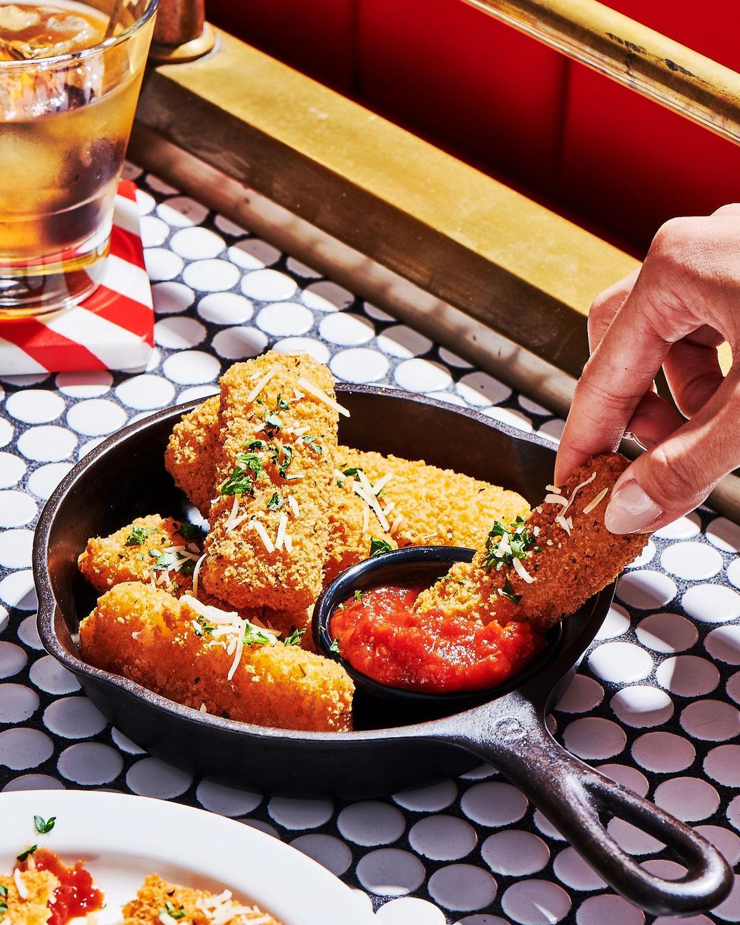 Crispy mozzarella sticks in cast iron skillet on mosaic tiled table with a red dipping sauce and hand reaching in to grab one.