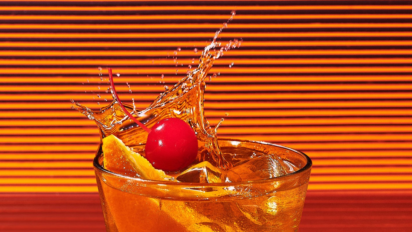 A glass of amber liquid with ice, garnished with an orange peel and cherry, shows a dynamic splash on an orange-striped backdrop.
