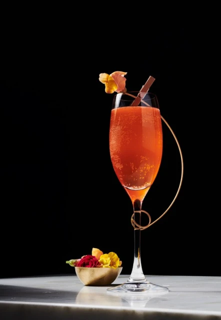 Elegant champagne flute filled with a sparkling orange cocktail, garnished with flowers and a chocolate stick, on a marble counter.