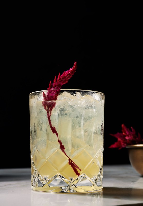 A cocktail in a faceted glass sits on a marble surface with a dark background, garnished with a red flower.