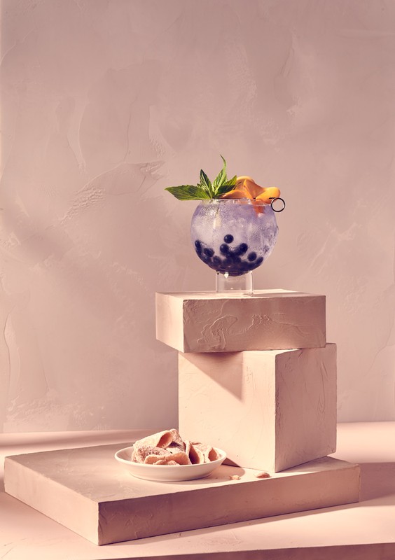 Cocktail in a round glass with blueberries, orange slice, and mint garnish, displayed on geometric blocks in a softly lit studio setting.