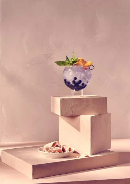 Cocktail in a round glass with blueberries, orange slice, and mint garnish, displayed on geometric blocks in a softly lit studio setting.