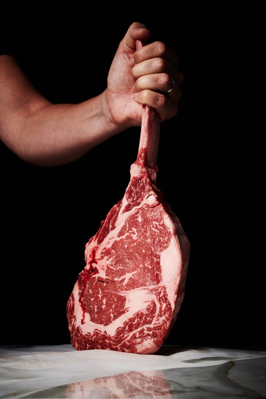 Close-up of a hand gripping a raw tomahawk steak against a dark background, highlighting the rich marbling and texture of the meat.