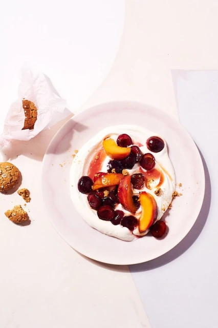 Overhead view of pavlova dessert on a plate with fresh cherries and peach slices, alongside cookies in a bright studio setting.