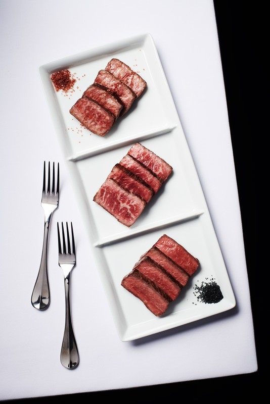 Overhead shot of sliced beef on a tiered white serving platter, accompanied by two forks and seasonings against a black backdrop.