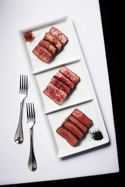 Overhead shot of sliced beef on a tiered white serving platter, accompanied by two forks and seasonings against a black backdrop.