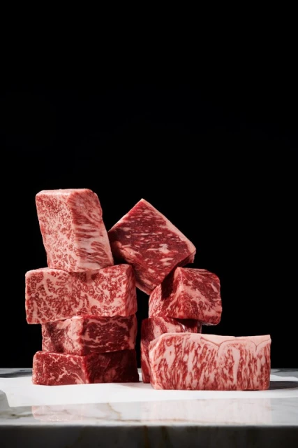 Wagyu beef cubes are stacked on a white surface against a black background, showcasing the meat's marbling and rich color.