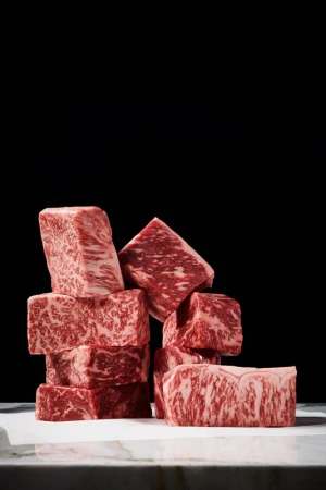Wagyu beef cubes are stacked on a white surface against a black background, showcasing the meat's marbling and rich color.