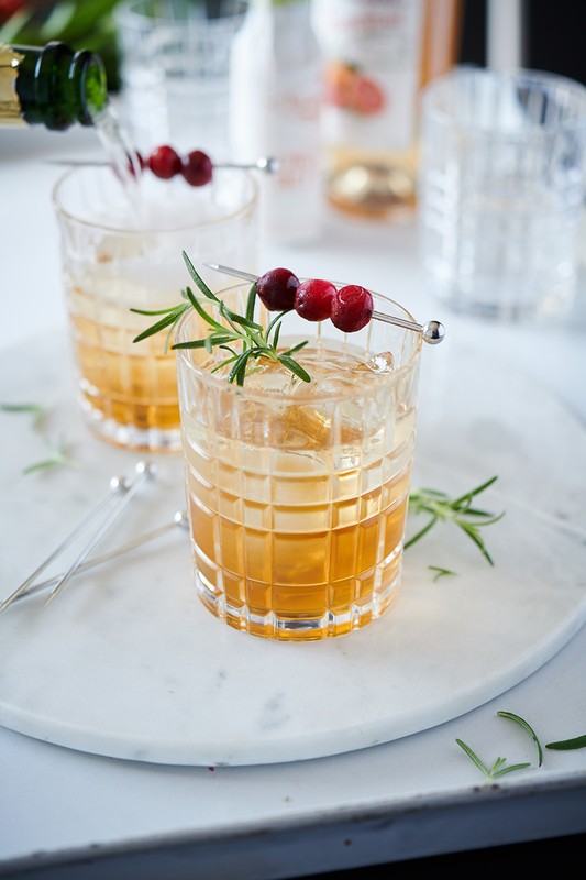 Two cocktails garnished with cranberries and rosemary sprigs sit on a marble tray, with bottles blurred in the background.