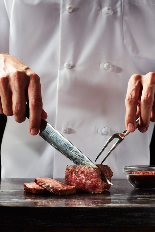 Close-up of chef cutting a steak with knife and fork on a dark countertop, showcasing culinary skill and the rich color of the meat.