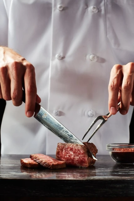 Close-up of chef cutting a steak with knife and fork on a dark countertop, showcasing culinary skill and the rich color of the meat.
