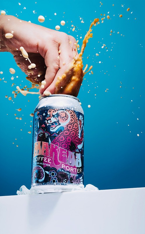 A hand splashes an open can of coffee porter. Liquid sprays in the air on a white surface in front of a bright blue backdrop.