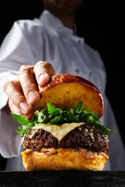 Close-up of a gourmet burger with a beef patty, melted cheese, and arugula, held by a chef in a white coat against a black background.