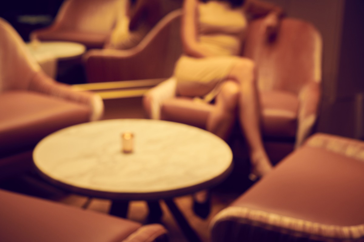 Blurred view of an adult female relaxing on pink chair near a round table with a candle in a dimly lit indoor lounge.