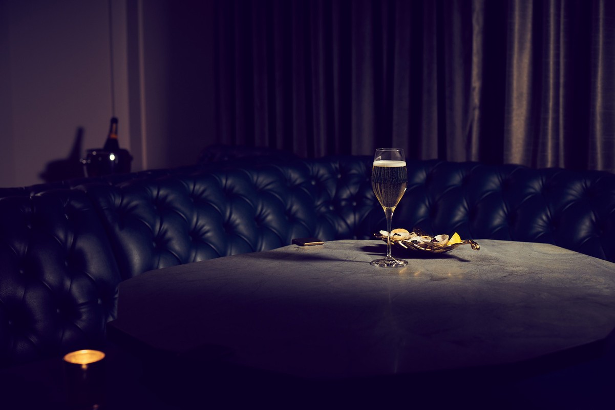 Champagne flute on a round table next to a leather tufted couch in a dimly lit room with a bottle in the background.