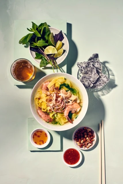 Overhead shot of Vietnamese wonton noodle soup with shrimp and pork, served with herbs, lime, dragon fruit, and dipping sauces.