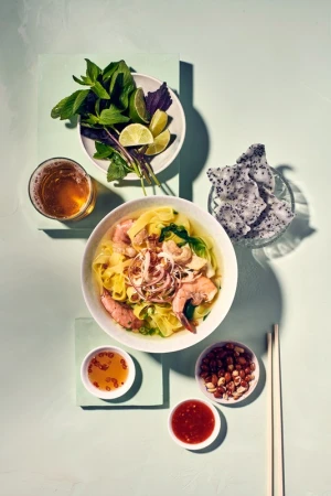 Overhead shot of Vietnamese wonton noodle soup with shrimp and pork, served with herbs, lime, dragon fruit, and dipping sauces.