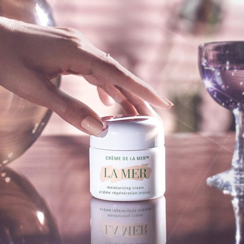 Close-up of a hand near a La Mer moisturizing cream jar on a reflective surface, alongside a tall glass; warm lighting.