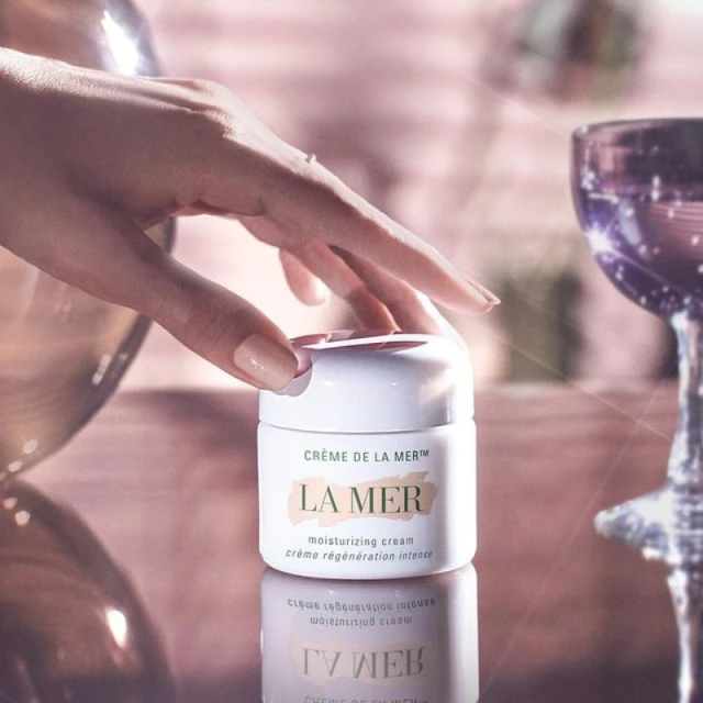 Close-up of a hand near a La Mer moisturizing cream jar on a reflective surface, alongside a tall glass; warm lighting.
