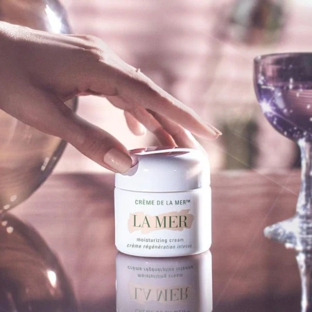 Close-up of a hand near a La Mer moisturizing cream jar on a reflective surface, alongside a tall glass; warm lighting.
