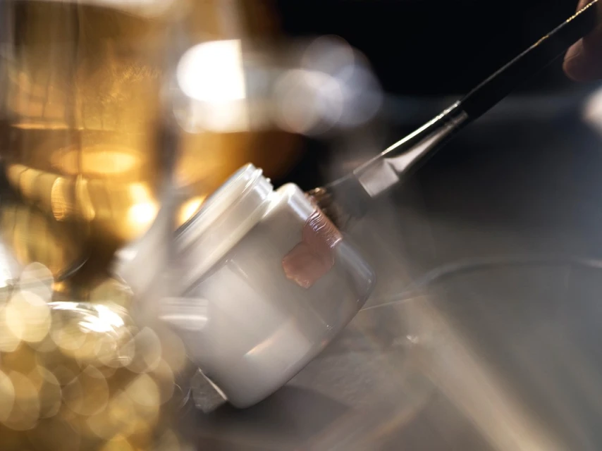 Close-up of a makeup brush applying cosmetic product from a jar in a blurred, brightly lit beauty setting.