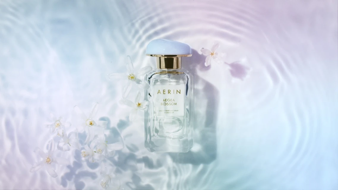AERIN Aegean Blossom perfume bottle on a light blue and purple water-like surface with white flowers, product shot for beauty industry.