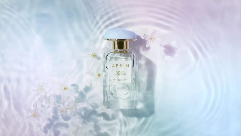 AERIN Aegean Blossom perfume bottle on a light blue and purple water-like surface with white flowers, product shot for beauty industry.