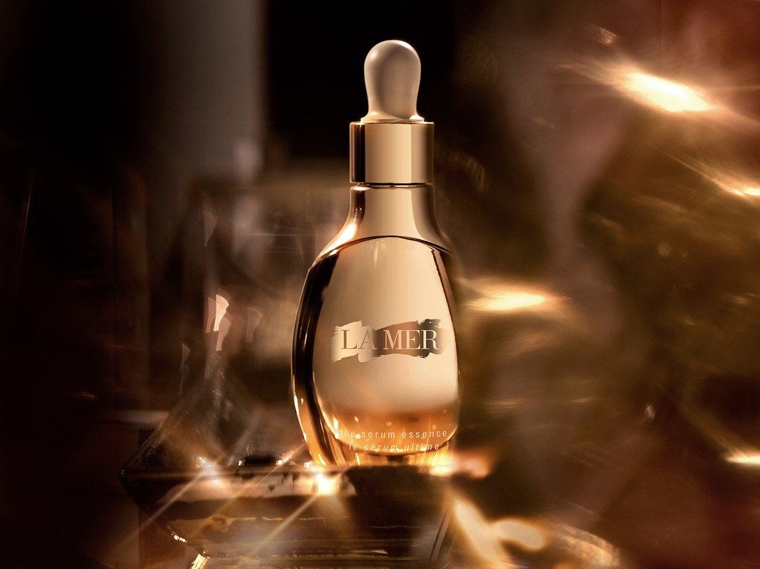 Close-up of gold La Mer serum bottle with white dropper on a dark background featuring golden bokeh and reflective surfaces.