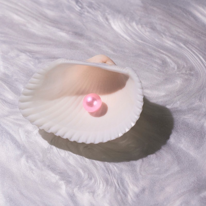 A pink pearl rests inside a white seashell, casting a soft shadow on a swirling, light-grey background, with beauty and elegance.