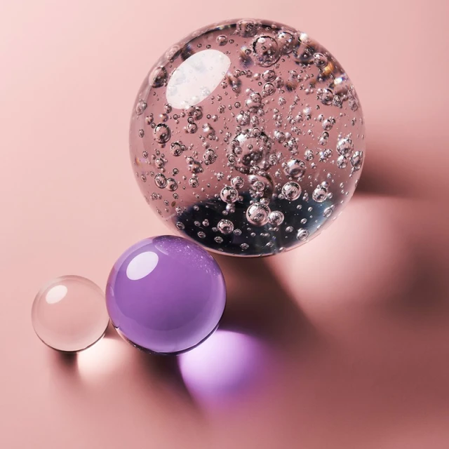 Three glass spheres of varying sizes and colors create an abstract still life on a solid pink background with cast shadows and reflections.