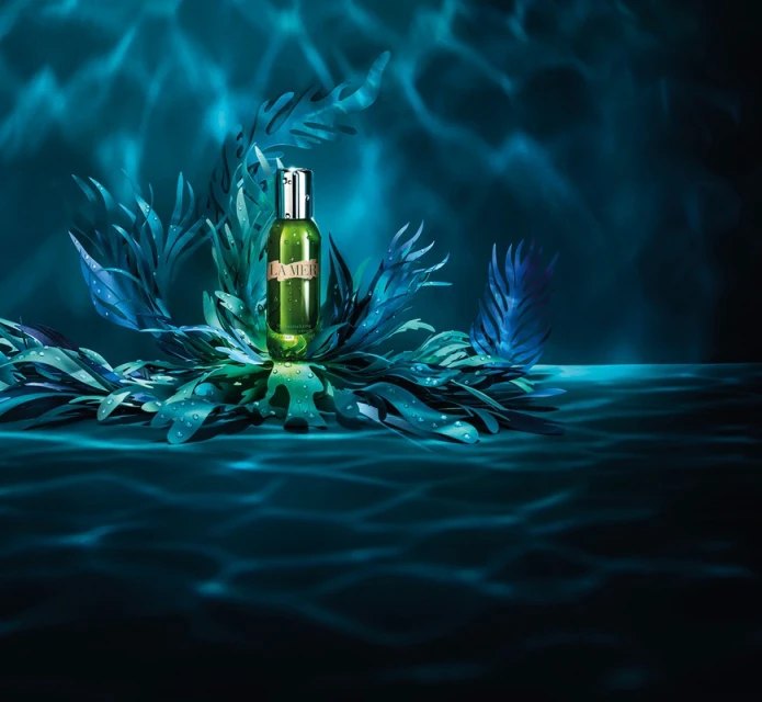 Product shot of a La Mer face oil bottle, arranged with faux seaweed fronds and featuring an underwater lighting effect and a blue backdrop.