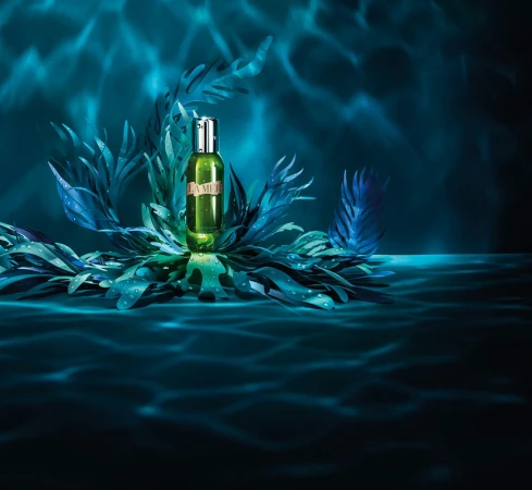 Product shot of a La Mer face oil bottle, arranged with faux seaweed fronds and featuring an underwater lighting effect and a blue backdrop.